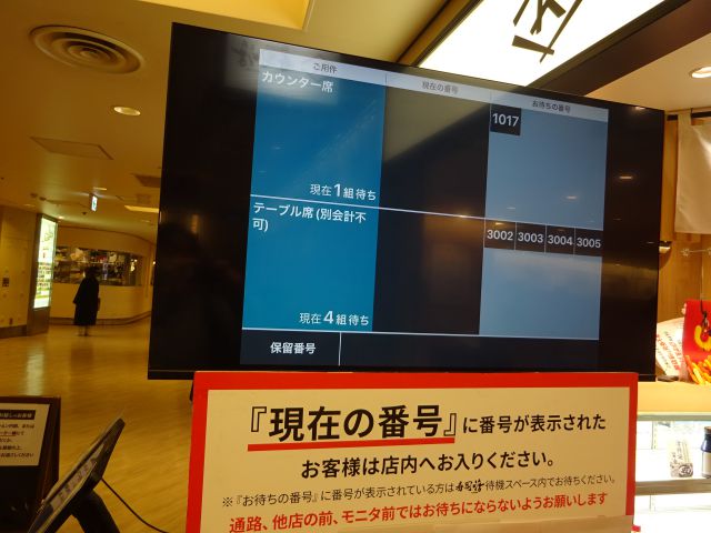 横浜駅西口「寿司活ジョイナス横浜店」の店頭に置かれた、順番待ちの番号が確認できる大型ディスプレイ（カウンター席とテーブル席それぞれ確認できる）